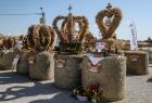 Widok na kilka wieńca dożynkowych, ustawionych na belach słomy. Wieniec na pierwszym planie wykonany jest w kształcie serca, zwieńcza go krzyż, pozostałe wieńce mają kształt koron również są zwieńczone krzyżem.