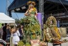 Widok na wieniec dożynkowy, który wykonany jest na kobiecym manekinie. Głowa figury przyozdobiona jest wiankiem z nawłoci. Suknia bogato zdobiona kwiatami i zielonymi liśćmi sięga jej do stup. 