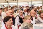 Kobiety i mężczyźni w strojach ludowych siedzą pod białym zadaszonym namiotem. Kobiety mają zawieszone na szyjach czerwone korale. 