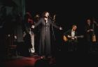 Dorota Ficoń aktorka Teatru Wiktacego podczas występu wokalnego, po lewej zespół aktorów teatru a po prawej zespół muzyczny
