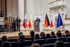 Widok na scenę filharmonii. Na której stoją Marszałek Witold Kozłowski i przedstawiciele władz samorządowych z Małopolski i Turyngii. Na scenie ustawiono flagę Polski, Małopolski, Niemczech i Unii Europejskiej. Przy pulpicie stoi przemawiający mężczyzna. 