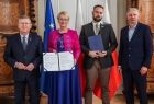 Marszałek Witold Kozłowski, Iwona Gibas z zarządu województwa, Michał Murzyn oraz mężczyzna stoją przy stole i trzymają egzemplarze podpisanych umów. W tle widoczne flagi: UE, Małopolski i Polski.