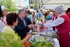 Marszałek Małopolski Witold Kozłowski, stoi przed jednym z białych stoisk, marszałek częstuję się regionalnymi produktami, które podaje mu kobieta ubrana w czerwony strój ludowy. 