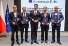 Wicemarszałek Łukasz Smółka stoi z beneficjentami na scenie. Z tyłu widoczne flagi Polski i Unii Europejskiej.