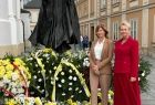 Iwona Gibas stoi obok pomnika Św. Jana Pawła II, przed pomnikiem widać wiązanki żółtych kwiatów przewiązane wstęgami