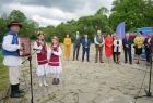 Ludzie w regionalnych strojach stoją przed mikrofonem. Z tyłu widoczni pozostali uczestnicy wydarzenia.