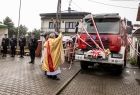 Ksiądz święci nowy wóz bojowy. Wóz strażacki przyozdobiony jest czerwonymi wstęgami. Z lewej strony widać stojących w strojach galowych strażaków ochotników. 