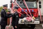 Wicemarszałek Łukasz Smółka wraz z innym strażakiem stoją na tle nowego wozu bojowego. Czerwony wóz strażacki, przepasany jest trzema biało- czerwonymi wstęgami. Marszałek Łukasz smółka odznacza stojącego przedni strażaka ochotnika. 
