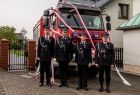Wicemarszałek Łukasz Smółka i trzech strażaków ochotników w strojach galowych. Mężczyźni stoją w rzędzie na tle czerwonego wozu bojowego. Wóz strażacki ozdobiony jest biało czerwonymi wstęgami. 
