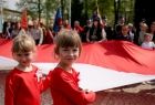 Dwie uśmiechnięte dziewczynki trzymające wraz z innymi osobami szeroko rozpostartą, trzymaną za krawędzie materiału biało-czerwoną flagę