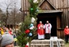 Dziewczynka z palmą wielkanocną. Z tyłu dwaj kapłani zwracający się do osób zebranych przed zabytkowym, drewnianym kościołem