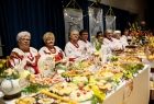 Długi stół wypełniony wielkanocnymi przysmakami, za nim na krzesłach ustawionych w rzędzie siedzą kobiety w strojach regionalnych