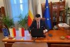 Wicemarszałek Łukasz Smółka w swoim gabinecie podczas konferencji online. W tle widoczne meble i flagi.