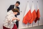 Wicemarszałek Łukasz Smółka pomaga wejść na scenę seniorce. Obok stoją biało-czerwone flagi.