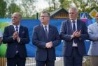 Marszałek Witold Kozłowski w towarzystwie dwóch mężczyzn. W tle widać niebieską baner zawieszony na płocie, dach drewnianego budynku i zielone drzewa.