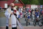 Z lewej strony zdjęcia stoi ksiądz, za nim widać kilkadziesiąt dzieci na rowerach, jadących ścieżką miasteczka rowerowego. W tle widać zabudowania domów i czerwone dachy. 
