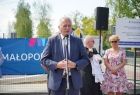 Mężczyzna w garniturze stoi przy mikrofonie, za nim stoi kilka osób, w tle widać niebieski baner z napisem Małopolska i logiem małopolska z lewej strony znajduję się biała tablica z informacją o funduszach pozyskanych na budowę miasteczka.