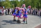 Dziewczynki w niebieskich strojach z różowymi pomponami stoją na środku placu, wokół nich zgromadziło się kilkadziesiąt osób. Cheerleaderki unoszą pompony nad głowy. W tle widać zielone drzewa i niebieskie niebo. 
