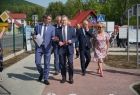 Wójt gminy Tymbark w otoczeniu samorządowców idzie środkiem ścieżki przy placu rowerowym. Z lewej strony widać zielony płot za którym znajduję się plac miasteczka rowerowego. W tle widać zabudowania i zielone drzewa. 