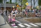 Alejki miasteczka pieszo rowerowego wypełnione dziećmi na rowerach i hulajnogach. Przy alejkach ustawiono znaki drogowe informujące o drodze z pierwszeństwem przejazdu, żółty znak ostrzegawczy, w tle widać drewniany budynek i zbudowania domów. 