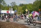 Alejki miasteczka rowerowego wypełnione rowerzystami. Widać dzieci na rowerach, hulajnogach i pieszych. Z lewej strony widać czerwony znak stopu i znak z pierwszeństwem przejazdu. W tle zielone drzewa.