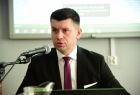 Wicemarszałek Łukasz Smółka przemawia do mikrofonu. W tle widoczny ekran.