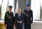 Marszałek Witold Kozłowski i dwóch strażaków w strojach galowych. Mężczyźni stoją na tle trzech flag Małopolskiej, Polskiej i Unijnej. 