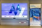 ekran z banerem promującym konferencję, obok rollup informującym o funduszach europejskich w Małopolsce