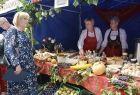 Na stole leżą spożywcze wyroby oraz warzywa i owoce. za stołem stoją dwie kobiety w ludowych strojach. Przed stołem stoi radna Marta Mordarska. 
