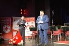 Poseł Patryk Wicher stoi na scenie w tle widać logo Polskiego Czerwonego krzyża. 