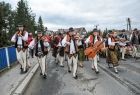 Bacowie ubrani w tradycyjne stroje z instrumentami muzycznymi m.in wiolonczela i skrzypcami idą środkiem drogi. Za nimi widać stado owiec. w tle widać zielone drzewa i miejskie zabudowania. 