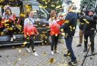 Trzy Cheerleaderki, w bluzach w kolorze biało czerwonym, dziewczynki wychodzą z czarnego busa, wokół nich widać złote konfetti.
