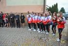 Grupa ludzi i cheerleaderki ustawieni są w półkolu, dziewczyny trzymają w rękach kwiaty, Z prawej strony zdjęcia widać ścianę budynku, z lewej samochody i zielone drzewa. 