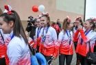 Cheerleaderki idą środkiem między kibicami, dziewczęta maja na sobie bluzy w kolorze białoczerwonym na niebieskiej wstążce maja zawieszone medale. Zawodniczki maja wpięte we włosy kokardy koloru biało czerwonego W tle widać grupę ludzi. 