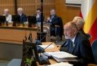 Sala Sejmiku Województwa Małopolskiego. Na pierwszym planie widać przewodniczącego prof. Jana Tadeusza Dude, pochylającego się nad mikrofonem. W tle widać flagę województwa małopolskiego.