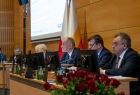 Na pierwszym planie członkowie prezydium sejmiku województwa małopolskiego. w prawym dolnym rogu widać bukiet czerwonych róż, w lewym górnym rogu znajduję się tablica multimedialna. Za prezydium znajduje się flaga w barwach małopolski. 