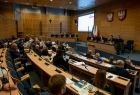 Widok z prawego boku sali sejmiku województwa małopolskiego. Radni wojewódzcy siedzą przy biurkach, w tle po lewej stronie widać tablice multimedialna, w Sali dominuje brązowy kolor ścian. 