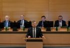 Marszałek Województwa Małopolskiego Witold Kozłowski, stoi przy pulpicie za nimi widać siedzących przy monitorach członków zarządu województwa małopolskiego. W sali sejmiku dominuję brązowy kolor ścian i biurek. 