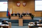 Marszałek Witold Kozłowski stoi przy pulpicie na środku sali sejmiku, za nim widać prezydium rady sejmiku. Po lewej i prawej stronie tyłem do zdjęcia siedzą radni wojewódzcy. 