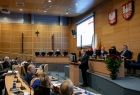 Widok z prawego boku sali Sejmiku Województwa Małopolskiego. Radni wojewódzcy siedzą przy biurkach ułożonych w lekkim łuku. W tle po lewej stronie widać tablice multimedialna, po niżej widać członków prezydium rady.