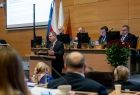 Widok na prezydium sejmiku województwa małopolskiego. Przy mównicy stoi marszałek Witold Kozłowski. W lewym górnym rogu widać fragment tablicy multimedialnej. Na pierwszym planie widać siedzących tyłem do zdjęcia radnych. 