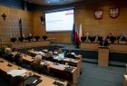 Widok z prawego boku sali Sejmiku Województwa Małopolskiego. Radni wojewódzcy siedzą przy biurkach ułożonych w lekkim łuku. w tle po lewej stronie widać tablice multimedialna, w sali dominuje brązowy kolor ścian.