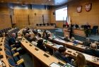 Widok z prawego boku sali Sejmiku Województwa Małopolskiego. Radni wojewódzcy siedzą przy biurkach ułożonych w lekkim łuku. w tle po lewej stronie widać tablice multimedialna, w Sali dominuje brązowy kolor ścian.