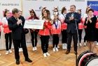 Wnętrze pomieszczenia, cheerleadereki ustawiły się obok siebie na tle białej ścianki z logami w kształcie litery M i napisem Małopolska. Marszałek Witold Kozłowski przekazuję symboliczne 100 % dofinansowanie na wyjazd reprezentacji.