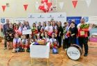 Wnętrze pomieszczenia, cheerleadereki ustawiły się w dwóch rzędach na tle białej ścianki z logami w kształcie litery M i napisem Małopolska. Przednimi ustawiono biały stolik z tortem. Z lewej strony widać grupę ludzi trzymających między innymi kwiaty.