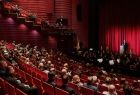 Widok na salę wypełniona ludźmi i scenę. Wewnątrz dominują ciemno czerwone kolory. 