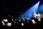 Widok na muzyków z filharmonii Warmińsko-Mazurskiej z Olsztyna w trakcie występów w Operze Krakowskiej. Fragment sceny na której stoją artyści jest podświetlony niebieskim punktowym światłem. 
