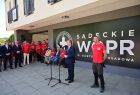 Marszałek Witold Kozłowski podczas wystąpienia, za nim stoją i siedzą na ławce ubrani w czerwone koszulki ratownicy WOPR