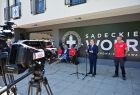 Marszałek Witold Kozłowski podczas wystąpienia, za nim stoją i siedzą na ławce ubrani w czerwone koszulki ratownicy WOPR; na pierwszym planie kamera telewizyjna