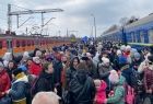 Uchodźcy z Ukrainy idą przez peron kolejowy. Po bokach widoczne pociągi.